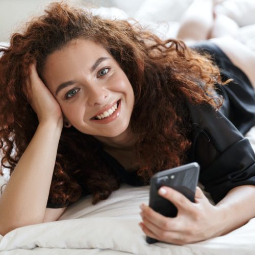 Happy cheerful positive woman in lingerie silk robe lies in home hotel on bed using mobile phone.